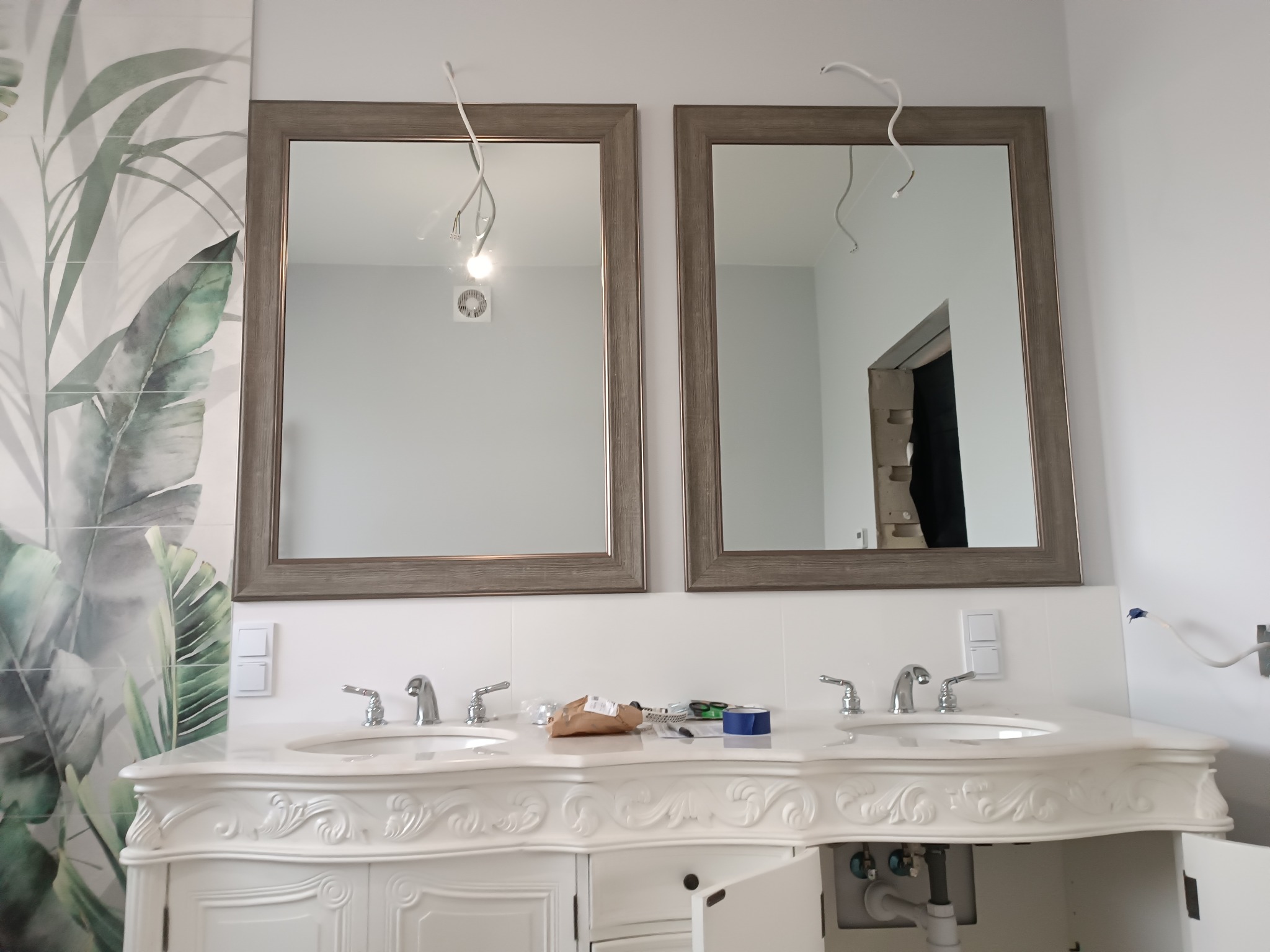 Biała, podwójna umywalka w stylu retro z ozdobnymi frezami, chromowanymi bateriami i dwoma lustrami w drewnianych ramach, widoczne niedokończone podłączenia elektryczne i narzędzia na blacie...