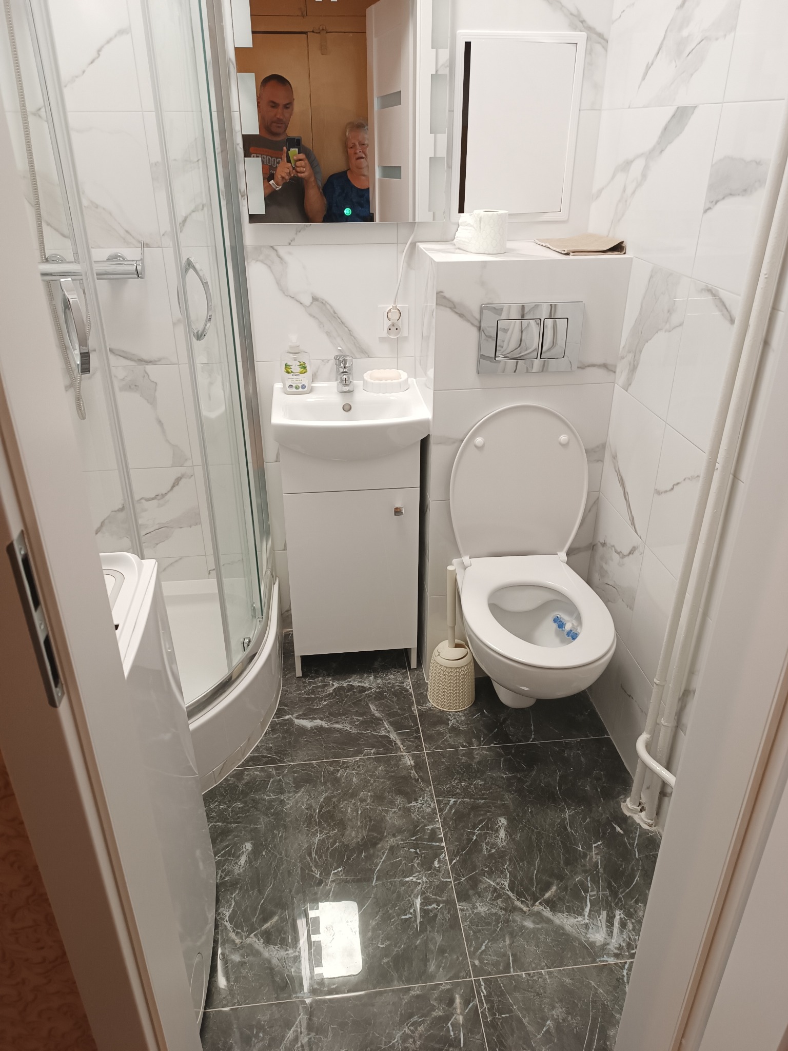 Nowoczesna, mała łazienka z kabiną prysznicową, umywalką z szafką, toaletą i marmurowymi płytkami na ścianach i podłodze. W lustrze odbija się osoba robiąca zdjęcie oraz starsza kobieta.