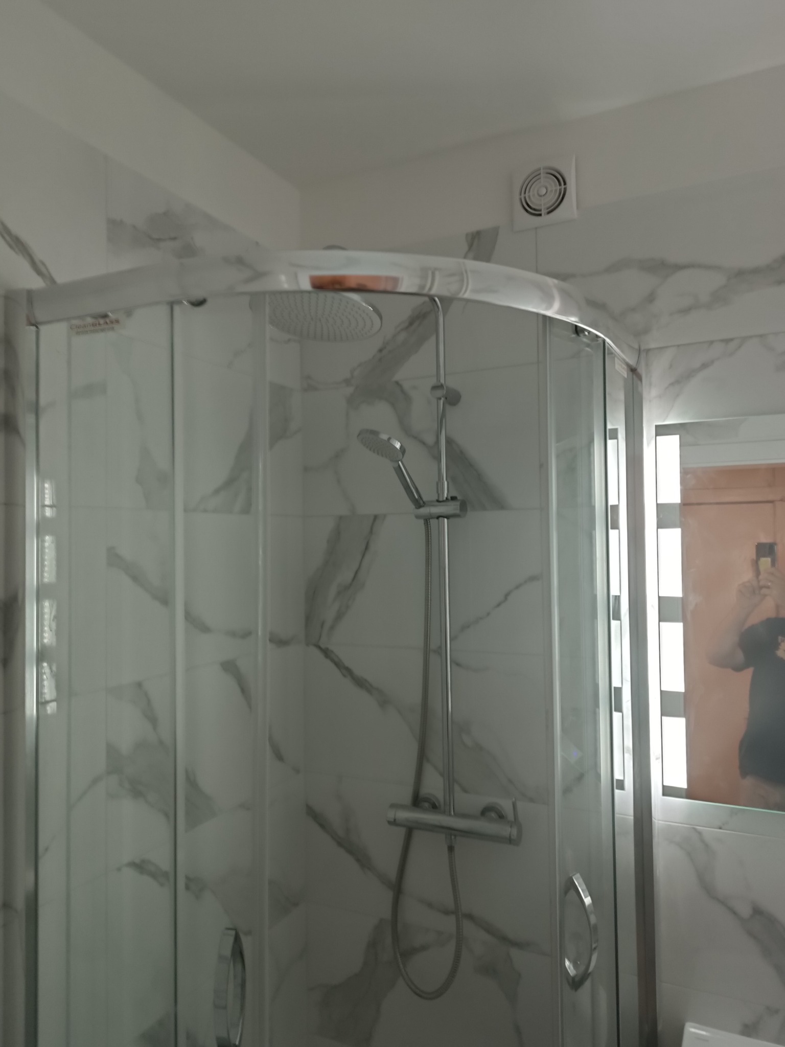 Nowoczesna kabina prysznicowa narożna z chromowanymi profilami i deszczownicą, wykończona płytkami imitującymi marmur, z widoczną kratką wentylacyjną na suficie i odbiciem osoby robiącej zdjęcie...