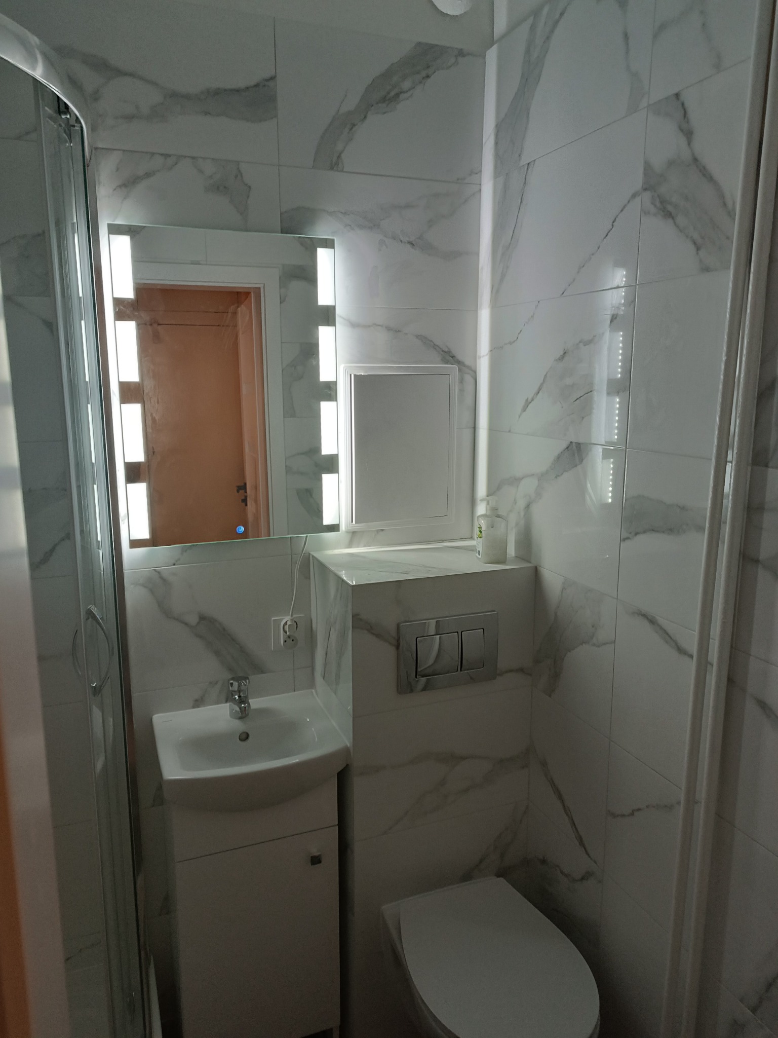 Nowoczesna, mała łazienka z białymi płytkami imitującymi marmur, wisząca umywalka z szafką, toaleta w zabudowie, podświetlane lustro i kabina prysznicowa ze szklanymi drzwiami.