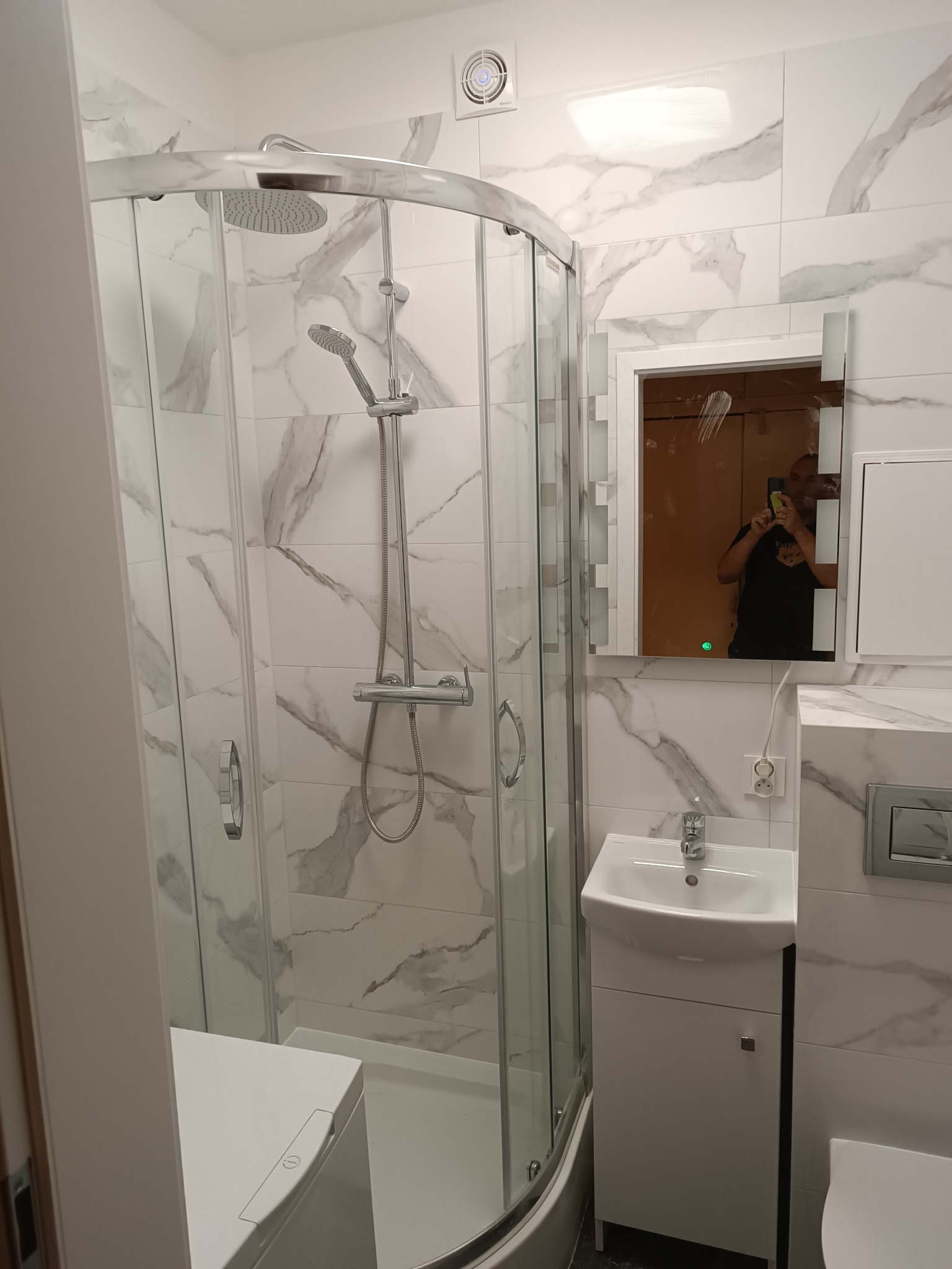 Nowoczesna łazienka z kabiną prysznicową typu walk-in z chromowanymi elementami, umywalką z szafką w kolorze białym oraz płytkami imitującymi marmur na ścianach; w lustrze odbija się osoba robiąca...