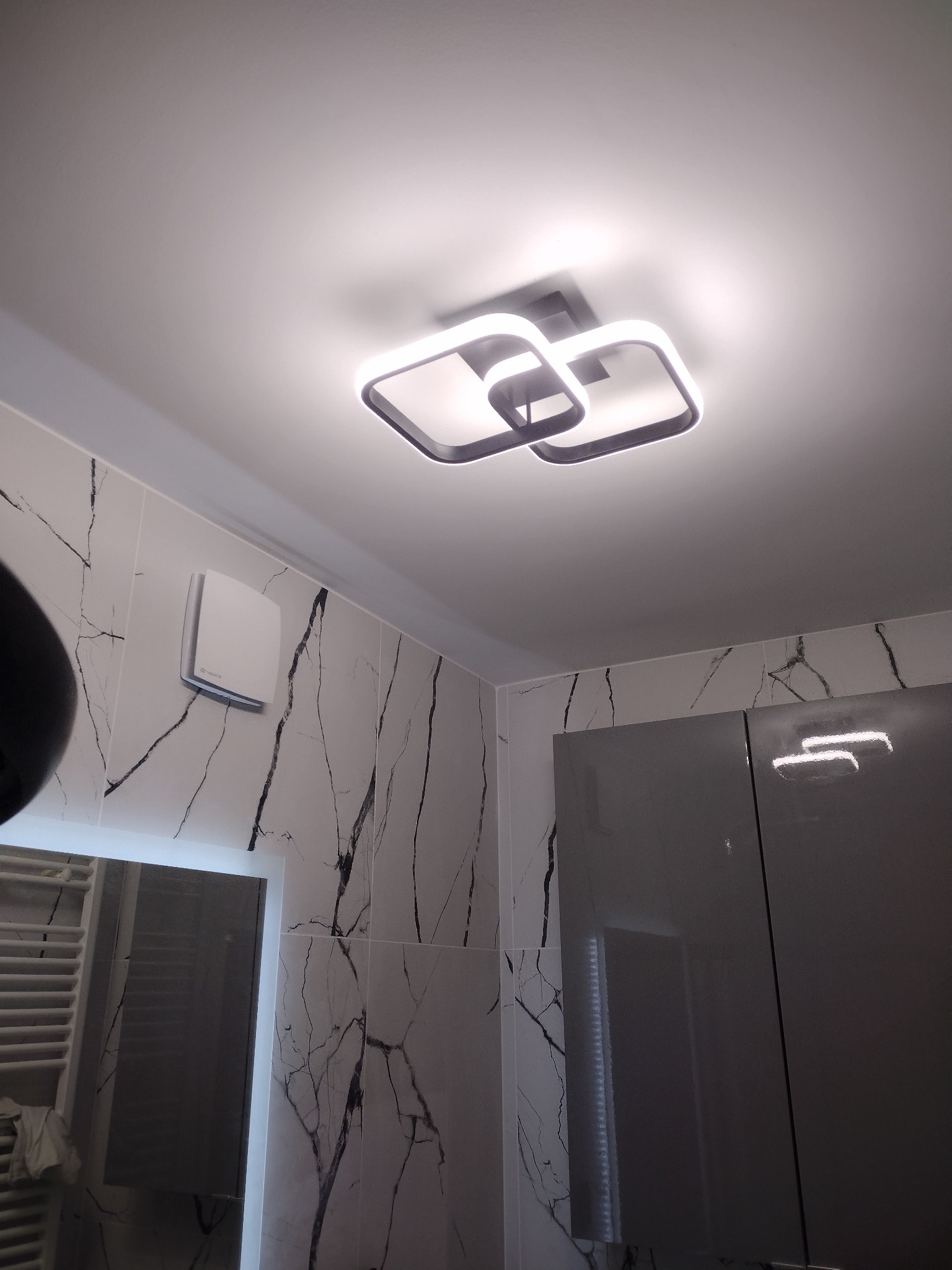 Nowoczesna lampa sufitowa LED o geometrycznym kształcie, zamontowana w łazience z marmurowymi płytkami i szarą zabudową meblową.