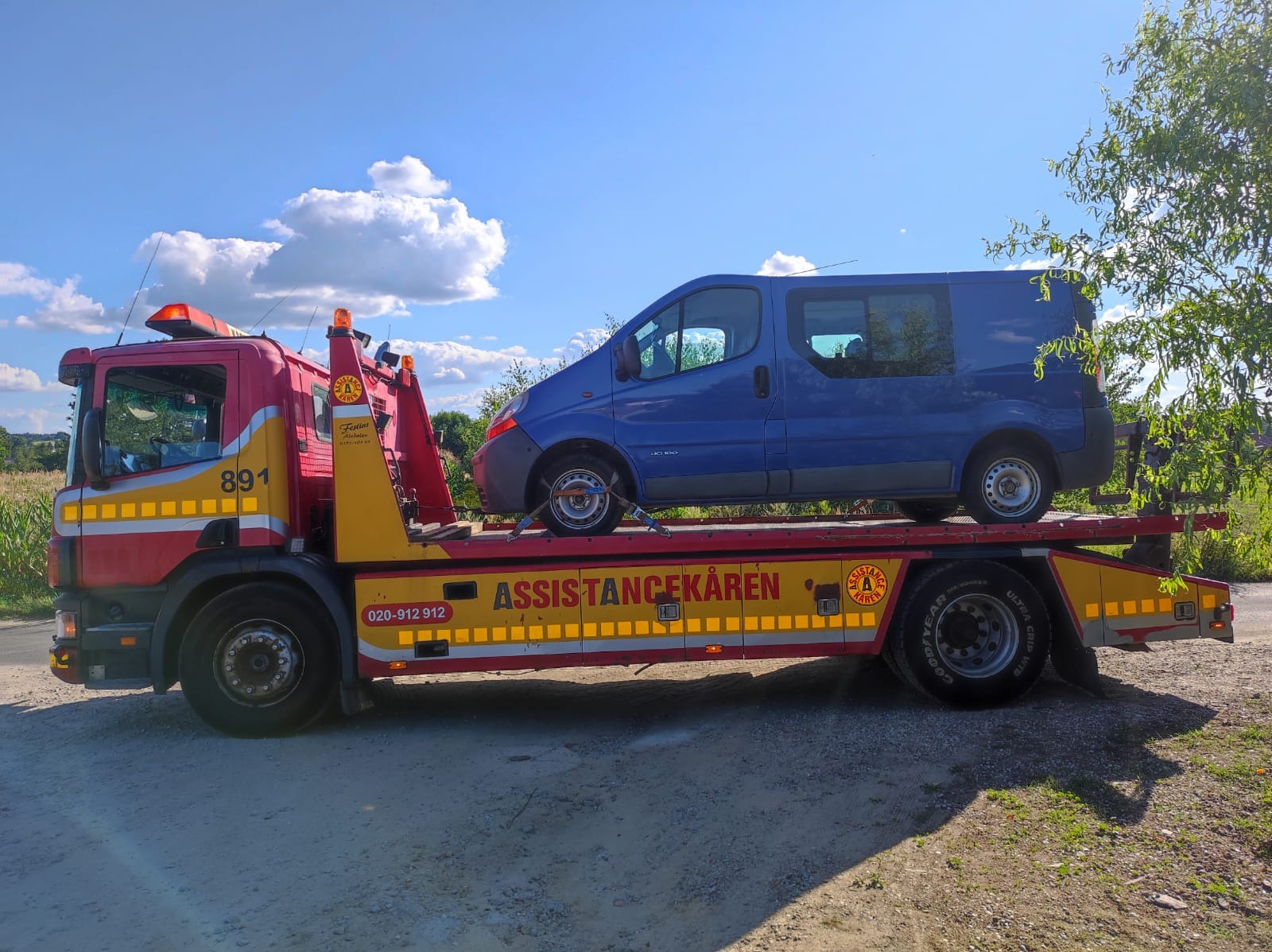 Czerwono-żółta laweta Assistancekåren z niebieskim busem Renault Trafic na platformie, zabezpieczonym pasami transportowymi, na szutrowej drodze w słoneczny dzień.