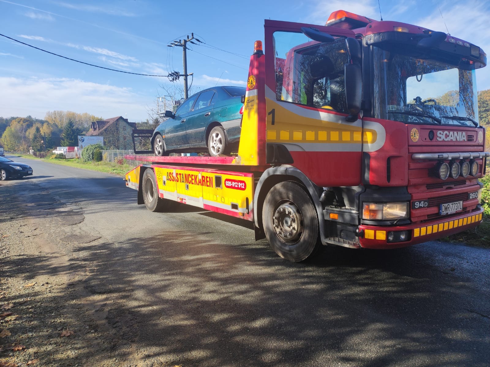 Czerwono-żółta laweta Scania z zielonym samochodem osobowym na platformie, widok z boku na drodze wiejskiej.