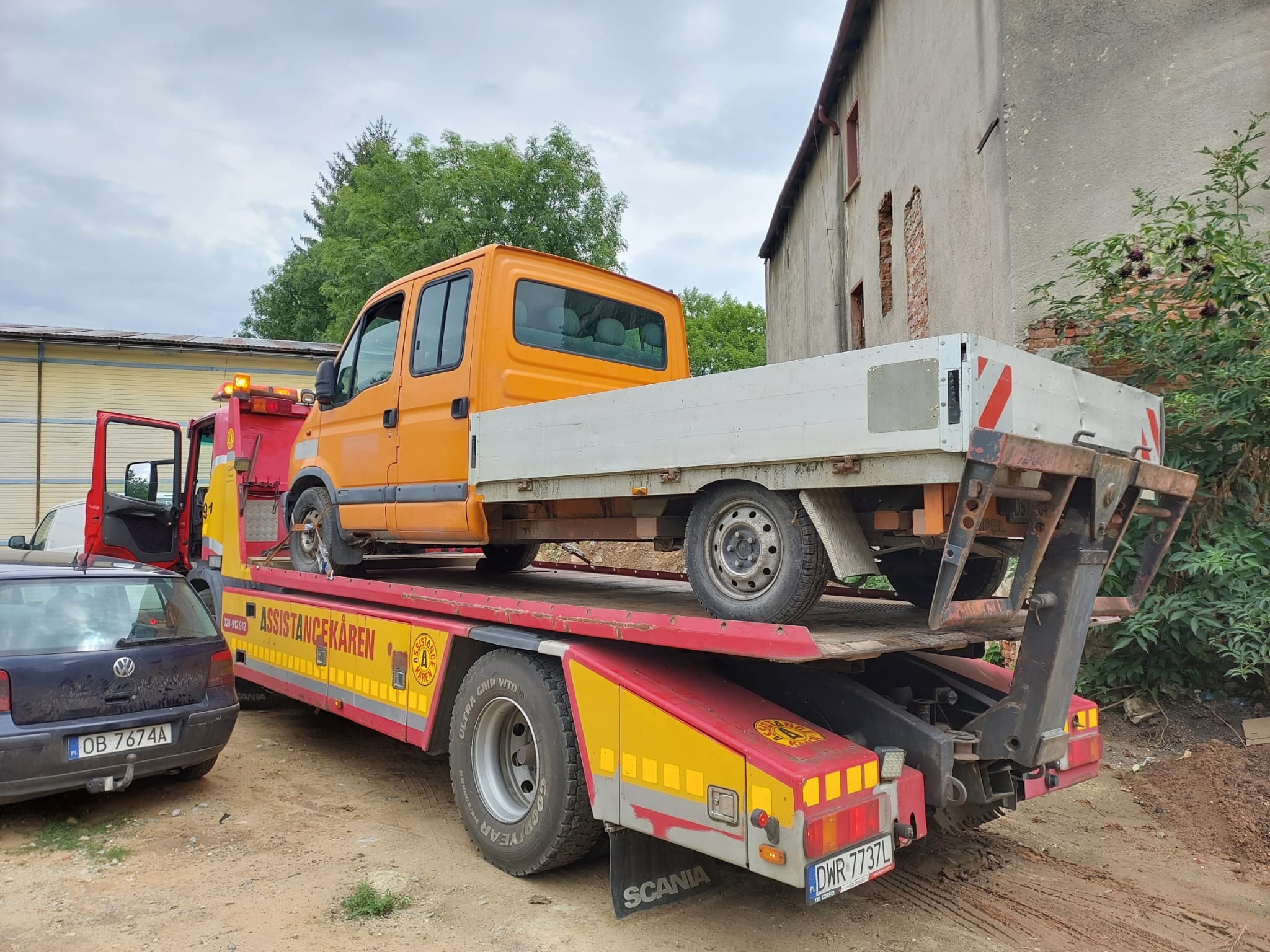 Pomarańczowy bus na czerwonej lawecie z napisem 'ASSISTANCEKAREN' na tle starego budynku i zaparkowanego samochodu osobowego.
