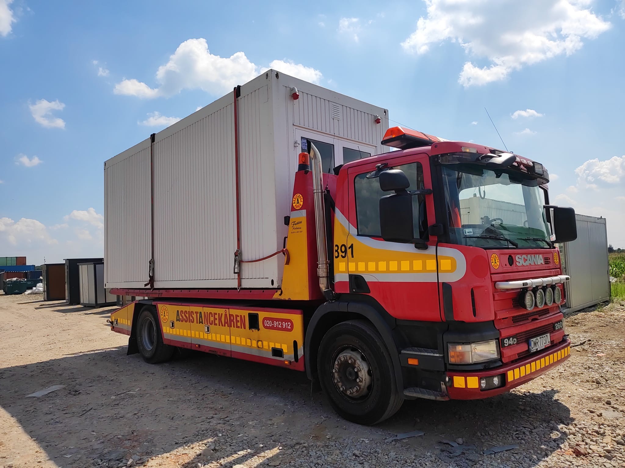 Czerwona laweta Scania z żółtymi elementami, przewożąca biały kontener modułowy z oknem, na żwirowym placu pod błękitnym niebem z chmurami.