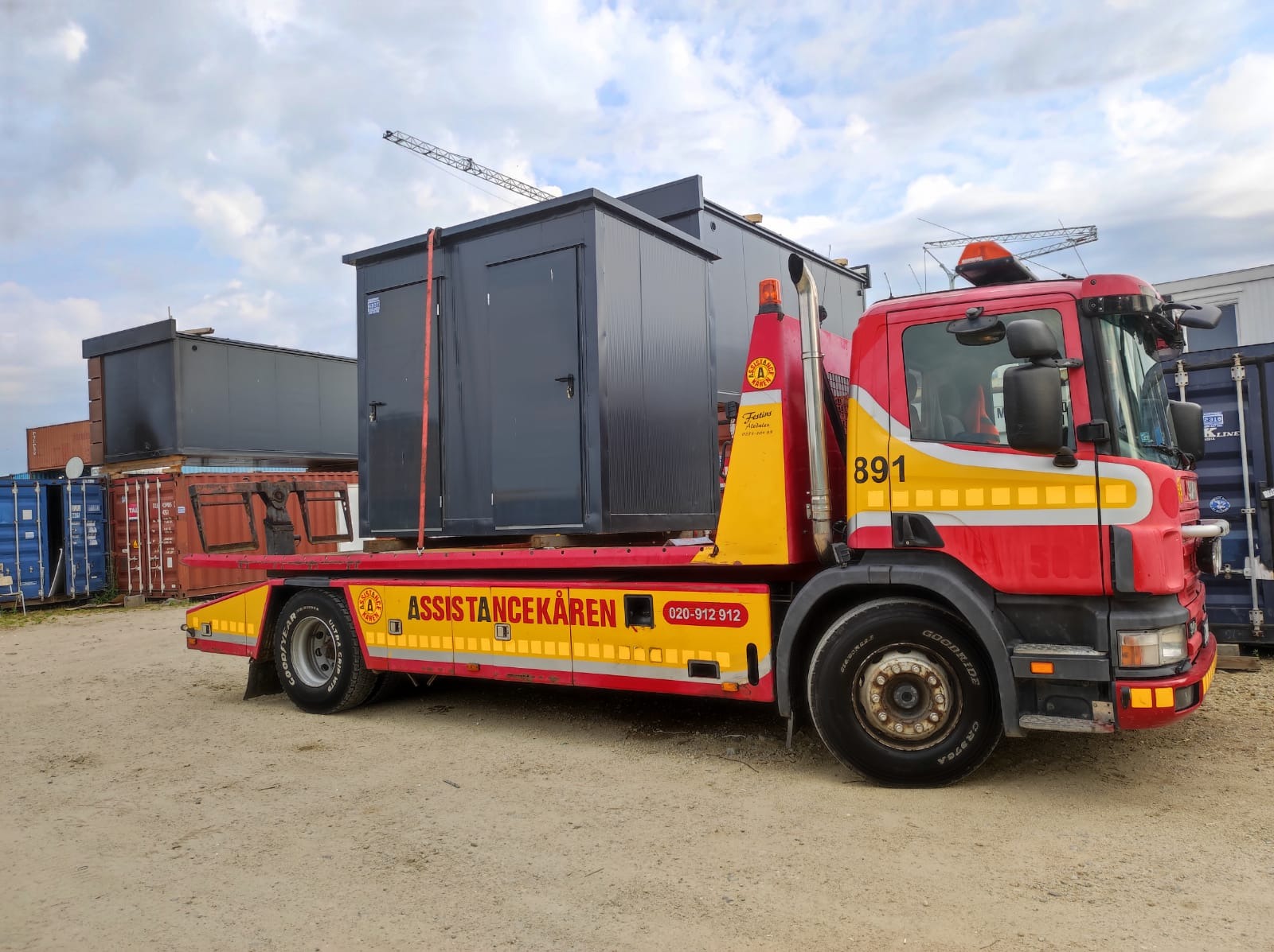 Czerwono-żółta laweta z załadowanym szarym kontenerem sanitarnym na tle placu z kontenerami i pochmurnego nieba.