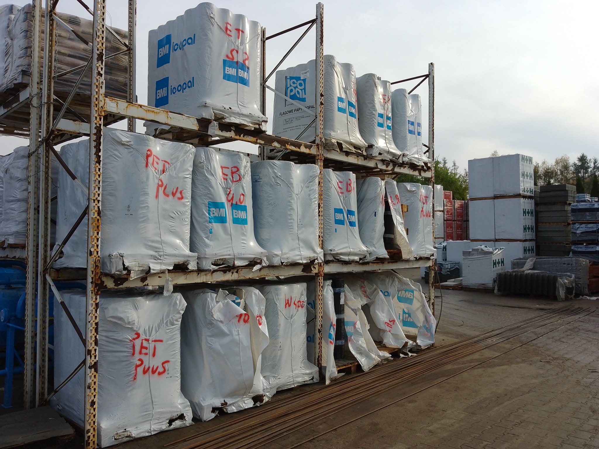 Magazyn materiałów budowlanych: rzędy zapakowanych rolek papy termozgrzewalnej na metalowych regałach, z napisem 'JET PLUS' i logo producenta, widoczne także inne materiały budowlane w tle.