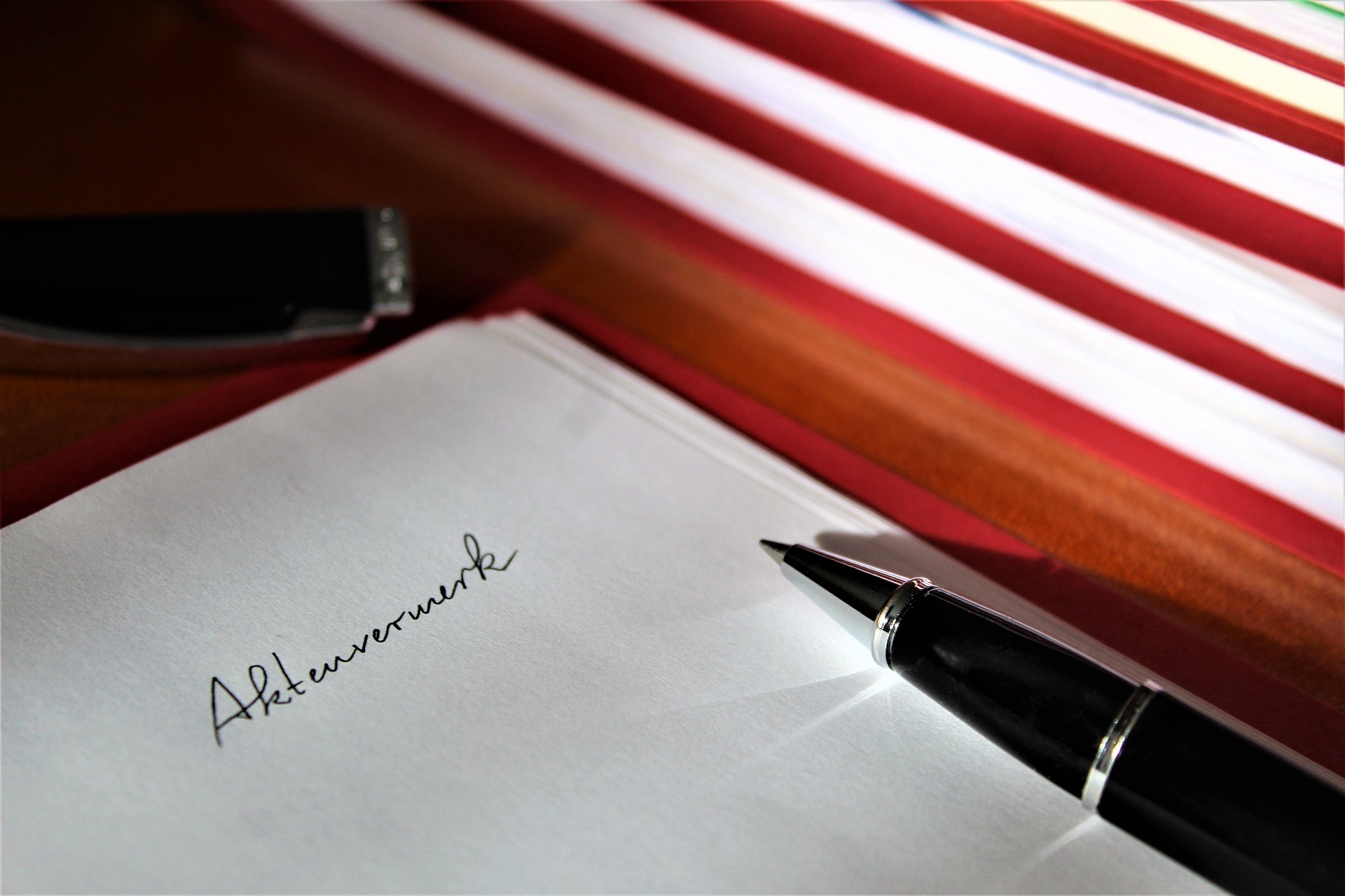 Czarny długopis na dokumencie z napisem 'Aktenvermerk', z otwartą skuwką i czerwono-białymi teczkami w tle. Stonowana kolorystyka, bliski kadr.