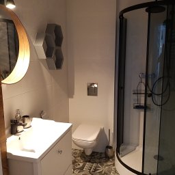 Bud-Mar - Nowoczesna łazienka w odcieniach bieli i szarości z kabiną prysznicową z czarnymi ramami, umywalką z szafką, toaletą i dekoracyjną półką w kształcie plastra miodu.