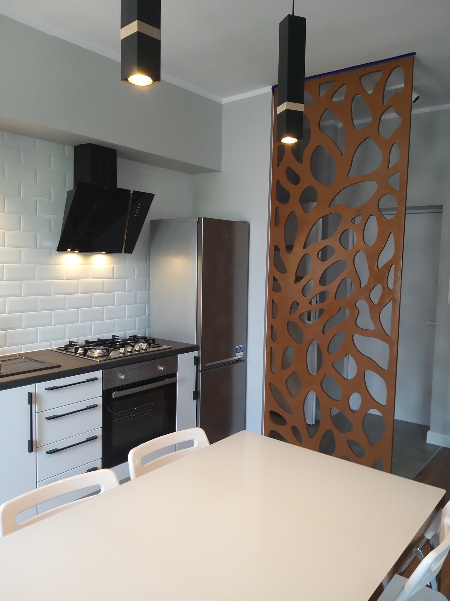 Nowoczesna kuchnia we Wrocławiu z białymi płytkami na ścianie, czarnym okapem i płytą gazową, oddzielona ażurowym, drewnianym panelem od części jadalnej z białym stołem i krzesłami.
