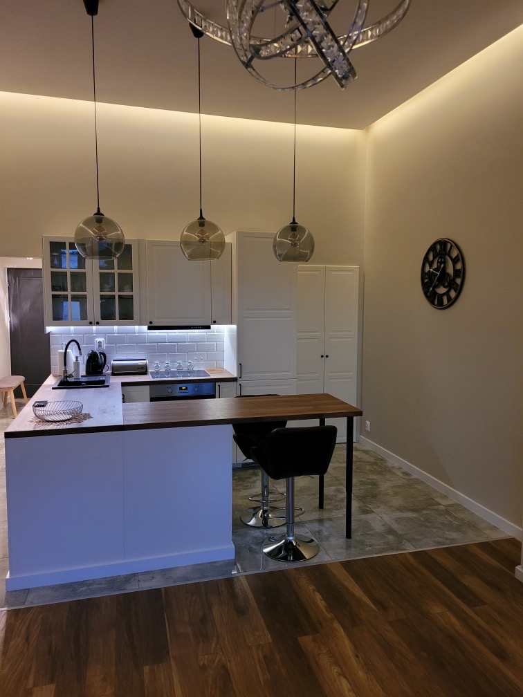 Nowoczesna kuchnia z białymi szafkami, wyspą z blatem w kolorze drewna i hokerami, oświetlona trzema szklanymi lampami wiszącymi i designerskim żyrandolem, z zegarem na ścianie.