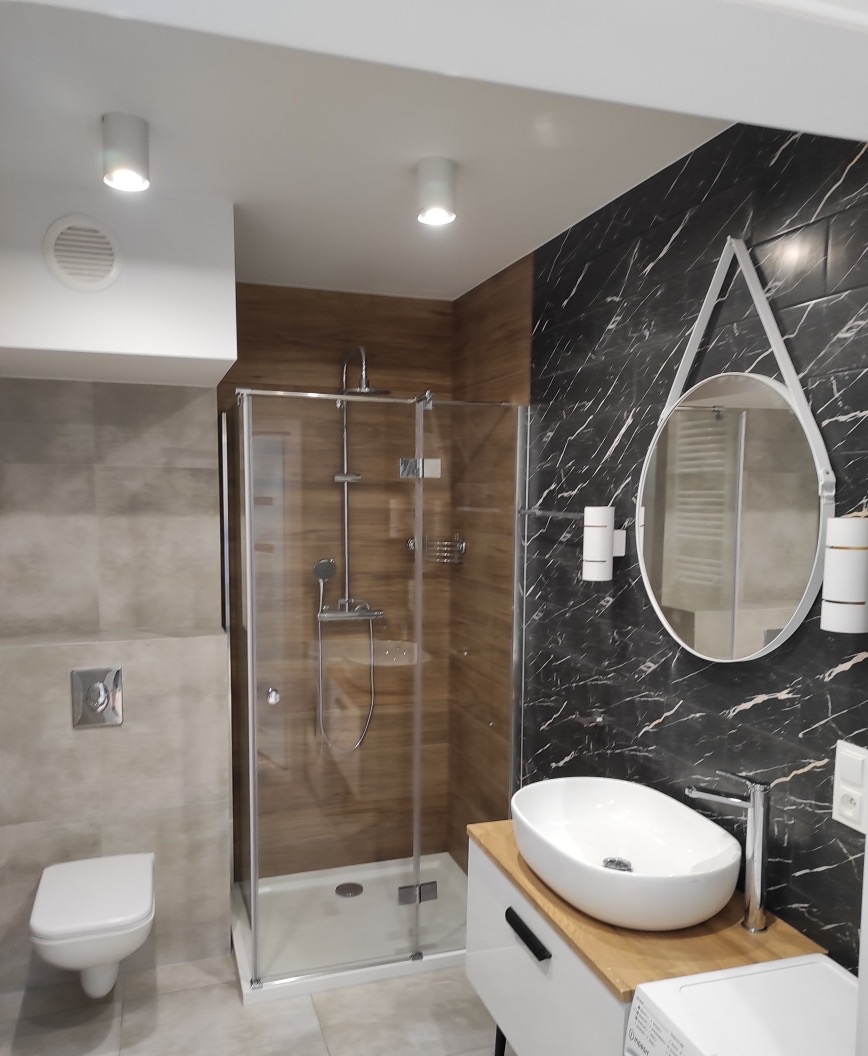Nowoczesna łazienka z kabiną prysznicową, umywalką nablatową na drewnianym blacie, okrągłym lustrem z białym obramowaniem i czarno-białą marmurową ścianą.