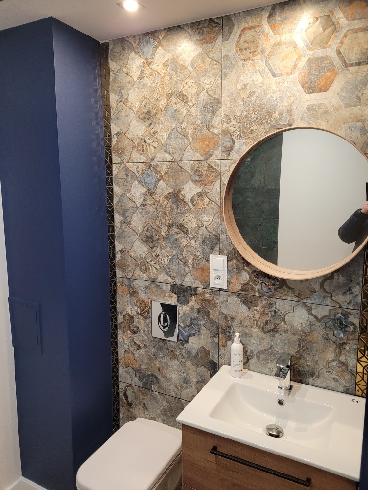 Wykończona łazienka z mozaikową glazurą w odcieniach brązu i szarości, okrągłe lustro z drewnianą ramą, biała umywalka z drewnianą szafką i armaturą oraz toaleta, na ścianie włącznik światła...