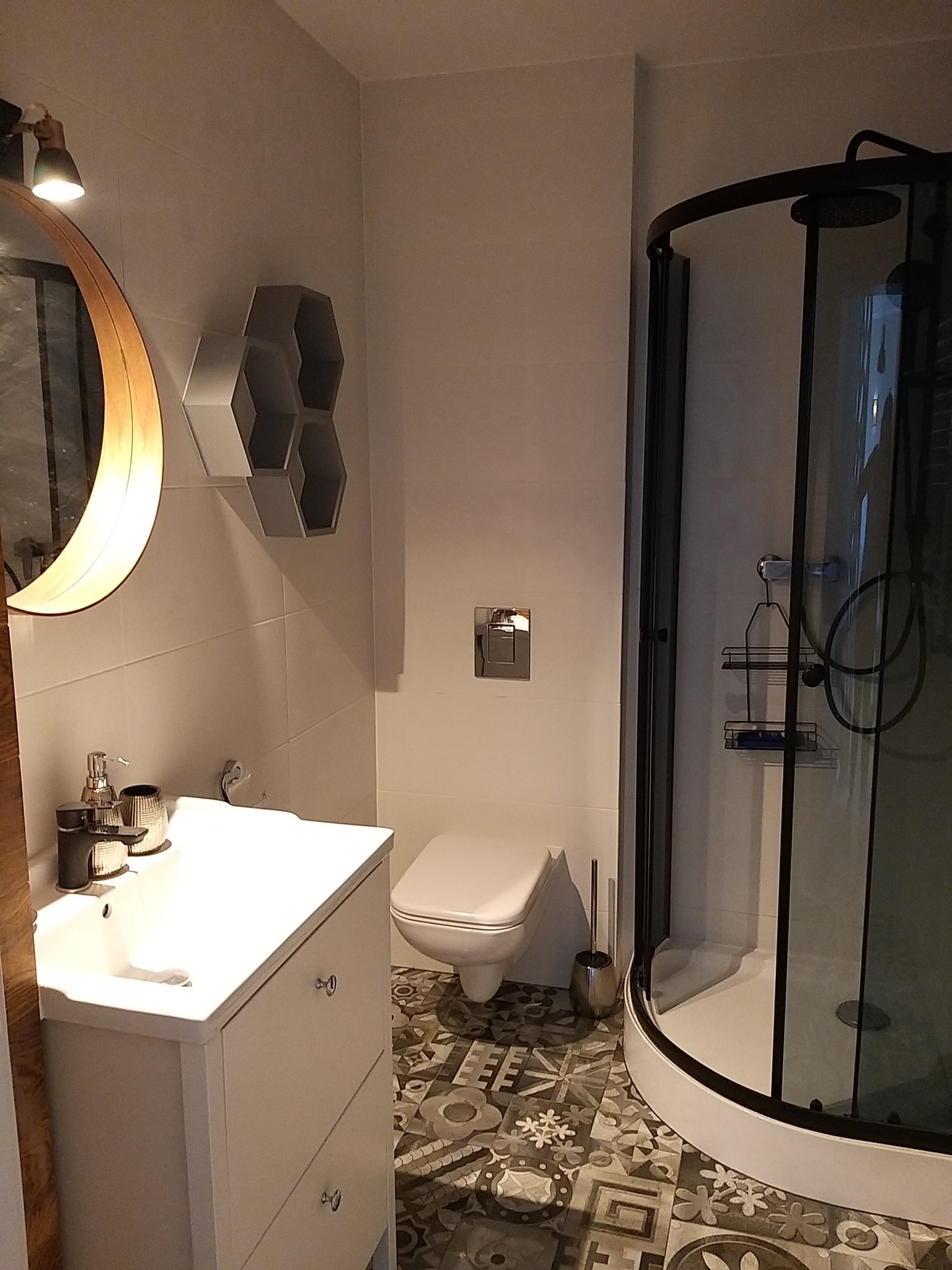 Nowoczesna łazienka w odcieniach bieli i szarości z kabiną prysznicową z czarnymi ramami, umywalką z szafką, toaletą i dekoracyjną półką w kształcie plastra miodu.