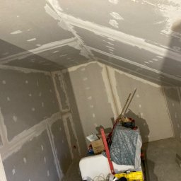 PERFECT_HOUSE - Poddasze w trakcie wykańczania płytami kartonowo-gipsowymi, widoczne łączenia płyt zaszpachlowane, na podłodze narzędzia i materiały budowlane, w tle ukośny sufit.