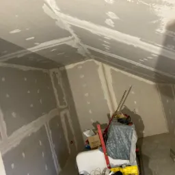 Poddasze w trakcie wykańczania płytami kartonowo-gipsowymi, widoczne łączenia płyt zaszpachlowane, na podłodze narzędzia i materiały budowlane, w tle ukośny sufit.
