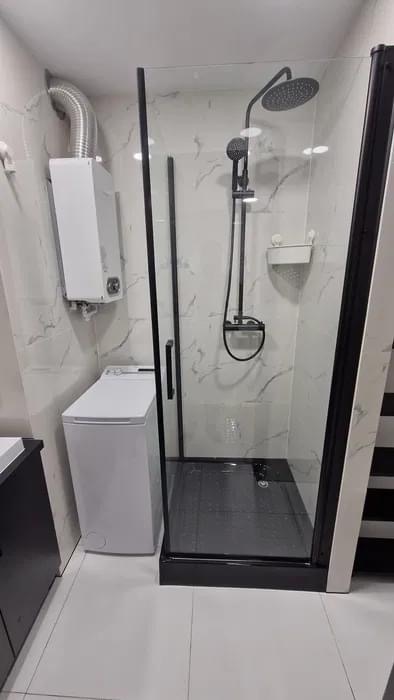Nowoczesna łazienka z kabiną prysznicową typu walk-in z czarnymi profilami, białą pralką i grzejnikiem wody na ścianie z marmurowym wzorem.