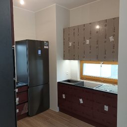 Northen - Kuchnia w trakcie montażu: widoczne szafki kuchenne z okleiną ochronną, jeszcze nie zdjętą, obok czarna lodówka, drewnopodobne płytki na podłodze, okno w tle.