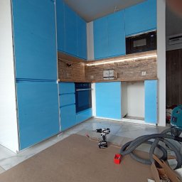 Northen - Niebieskie szafki kuchenne z białymi krawędziami, częściowo zamontowane, z widoczną ceglaną ścianą nad blatem, oświetleniem LED i narzędziami na podłodze.