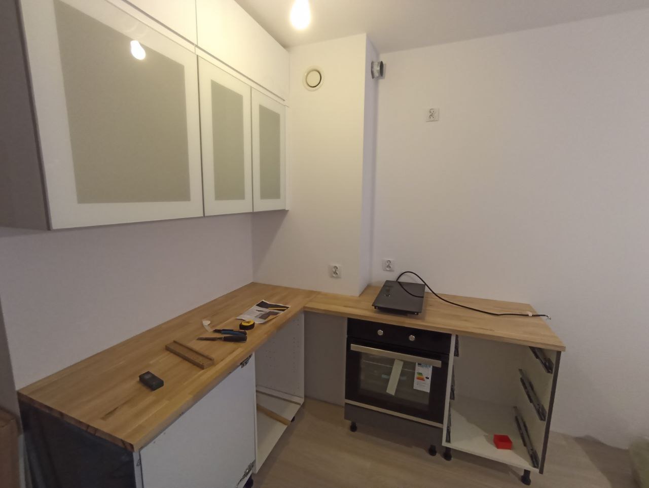 Fragment kuchni w trakcie montażu: drewniany blat roboczy, częściowo zamontowane szafki wiszące z mlecznymi szybami, płyta indukcyjna na blacie obok piekarnika, narzędzia i listwy na blacie.