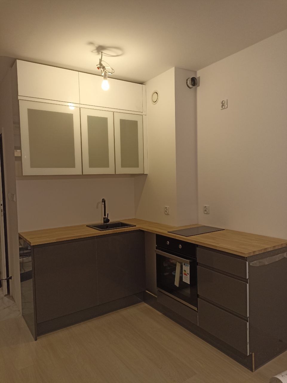 Nowoczesna kuchnia w odcieniach szarości z drewnianym blatem, czarnym zlewem i płytą indukcyjną, oświetlona pojedynczą żarówką.