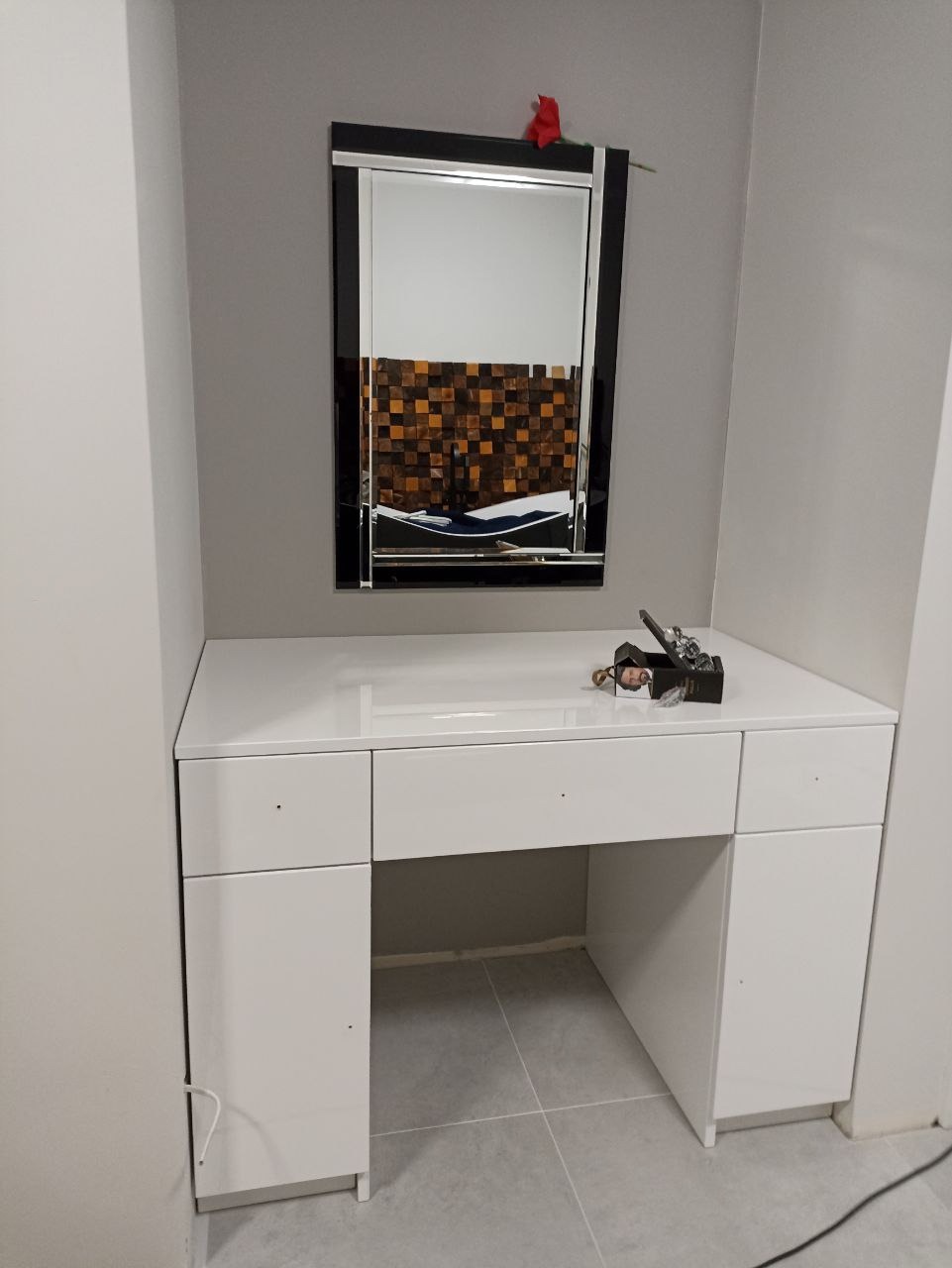 Biała toaletka z lustrem w czarnej ramie i czerwoną różą na górze, stojąca na szarych płytkach podłogowych.