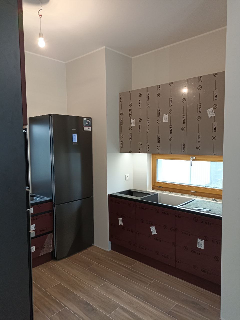 Kuchnia w trakcie montażu: widoczne szafki kuchenne z okleiną ochronną, jeszcze nie zdjętą, obok czarna lodówka, drewnopodobne płytki na podłodze, okno w tle.