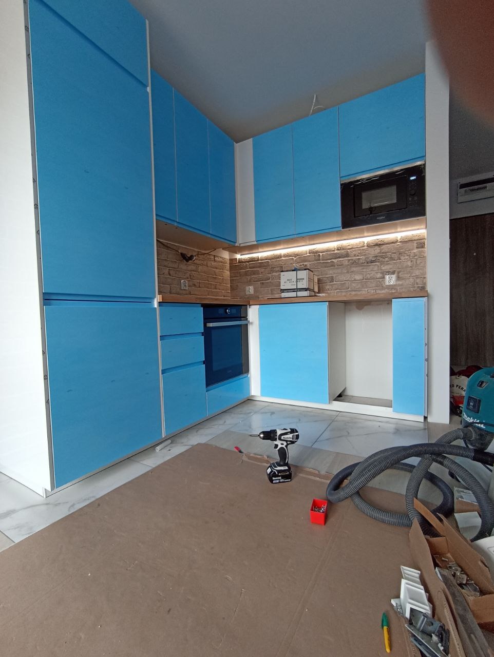 Niebieskie szafki kuchenne z białymi krawędziami, częściowo zamontowane, z widoczną ceglaną ścianą nad blatem, oświetleniem LED i narzędziami na podłodze.