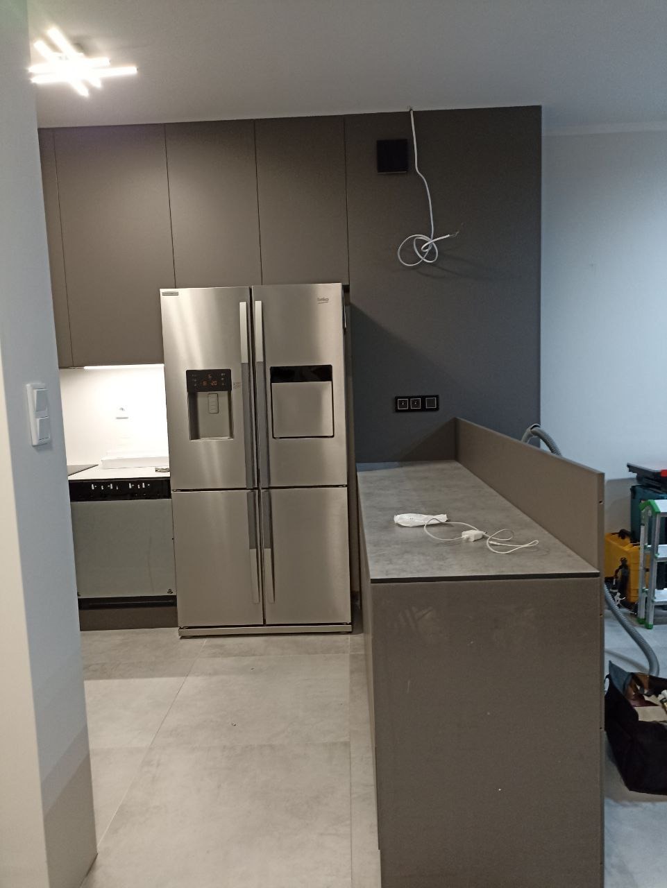 Szara kuchnia z zabudową, srebrną lodówką side-by-side i niedokończonymi instalacjami elektrycznymi.