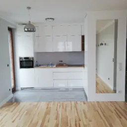 Nowoczesna, jasna kuchnia z białymi szafkami i drewnianą podłogą ułożoną w jodełkę, widok z przodu na część pomieszczenia z lustrem wbudowanym w ścianę.