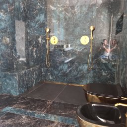 Mieczkowski Budownictwo - Luksusowa łazienka z ciemnymi marmurowymi płytkami, złotą armaturą i kabiną prysznicową typu walk-in. W lustrzanej powierzchni odbija się osoba robiąca zdjęcie.