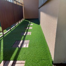 Mieczkowski Budownictwo - Ułożona sztuczna trawa na balkonie z drewnianymi płytkami tworzącymi ścieżkę, widok z góry, cień rzucany przez balustradę