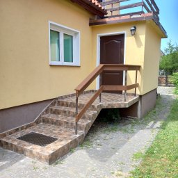 Mieczkowski Budownictwo - Żółty budynek z brązowymi drzwiami i schodami wyłożonymi brązowymi płytkami z metalową kratką ściekową, drewniana balustrada, widok z zewnątrz.