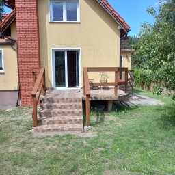 Mieczkowski Budownictwo - Żółty dom z ceglanym kominem i tarasem wyłożonym płytkami, z brązową drewnianą balustradą i schodami, otwartymi drzwiami balkonowymi i krzesłem na tarasie.