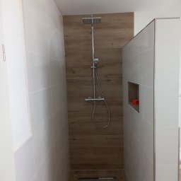 Mieczkowski Budownictwo - Nowoczesny prysznic typu walk-in z drewnopodobną ścianą i białymi płytkami, widoczny odpływ liniowy w podłodze.