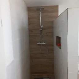 Nowoczesny prysznic typu walk-in z drewnopodobną ścianą i białymi płytkami, widoczny odpływ liniowy w podłodze.