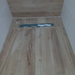 Płytki podłogowe imitujące drewno w kabinie prysznicowej z odpływem liniowym, jasne fugi.