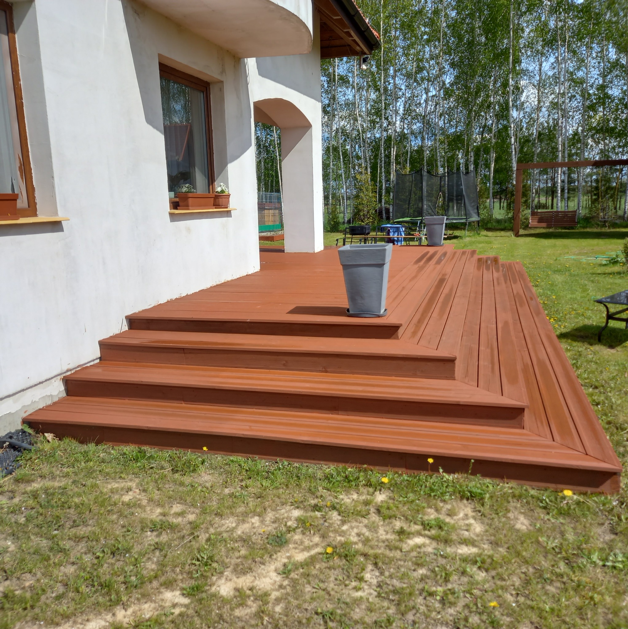 Nowo wybudowany drewniany taras z schodami, pomalowany na brązowo, z szarą donicą na tle domu z białymi ścianami i zielonego trawnika z drzewami w tle.
