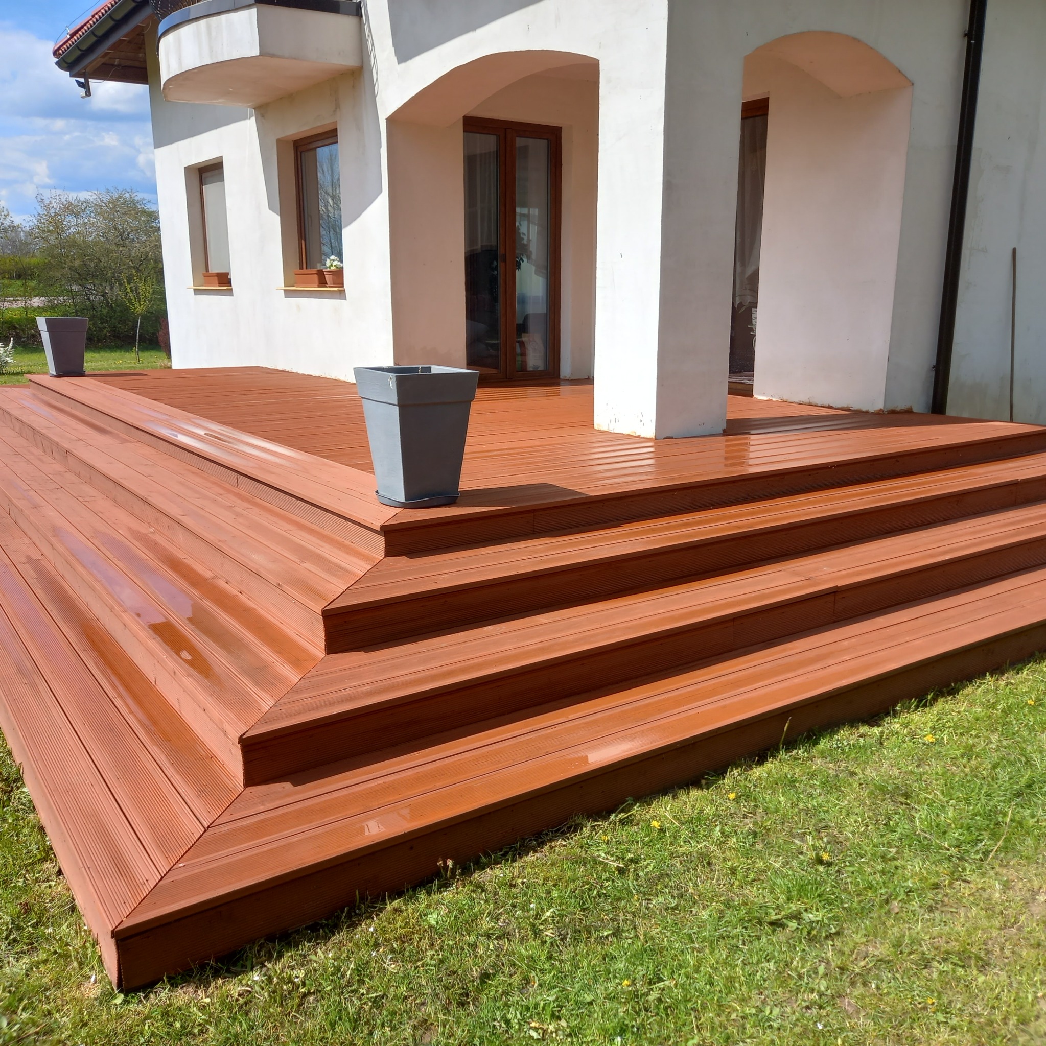 Nowy, brązowy taras z desek kompozytowych prowadzący do wejścia w białym budynku, z donicami po bokach.