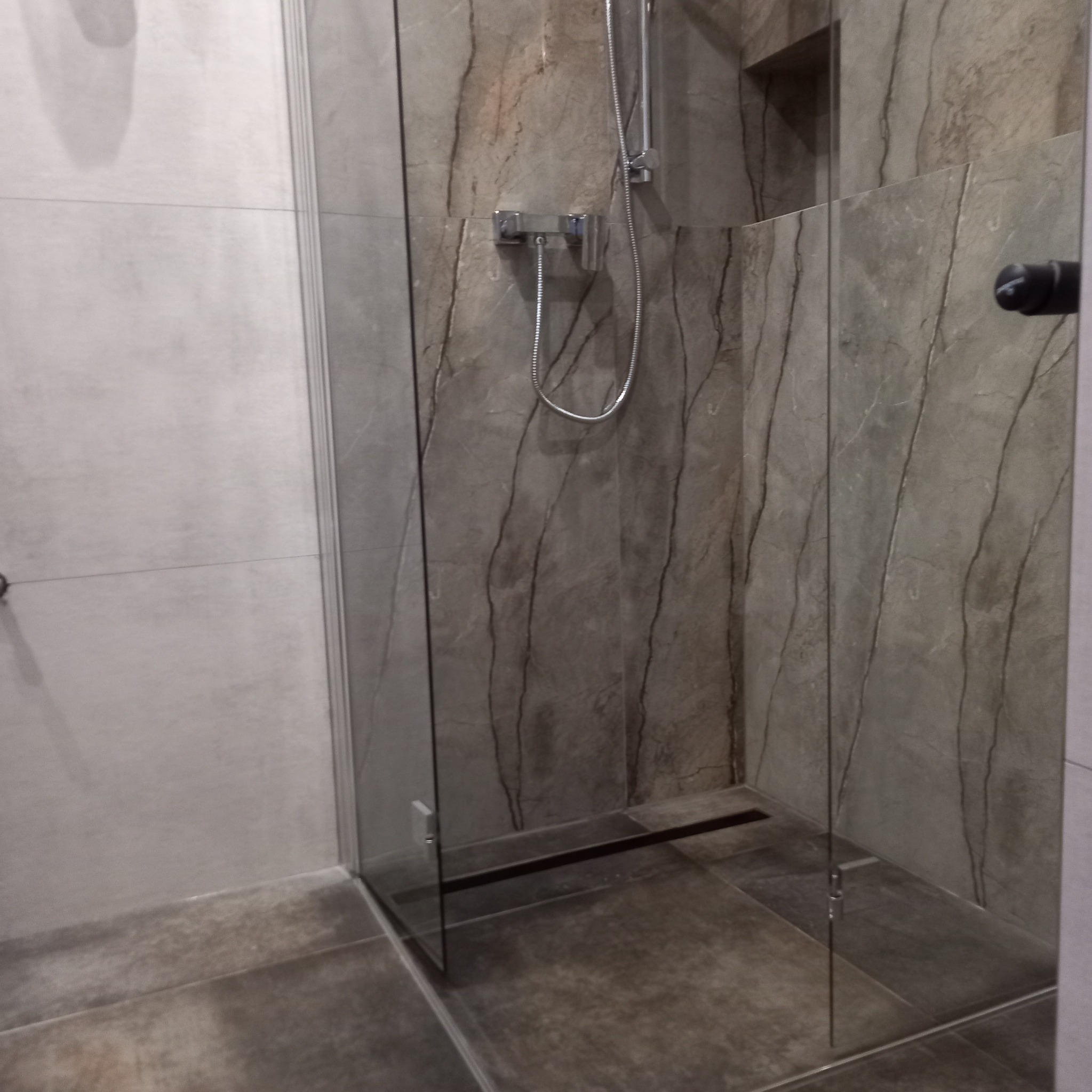 Nowoczesna kabina prysznicowa walk-in z szarymi płytkami imitującymi marmur na ścianach i podłodze, chromowana bateria i deszczownica, widoczny odpływ liniowy.