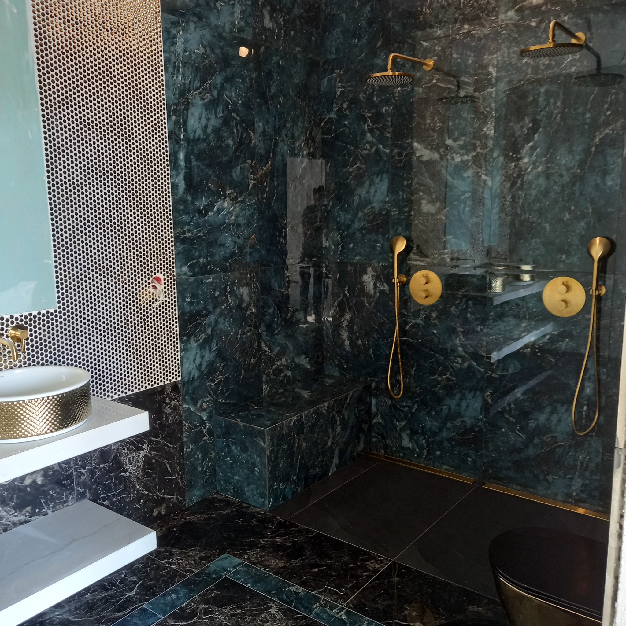 Luksusowa łazienka z zielonymi marmurowymi ścianami, złotymi bateriami i deszczownicami, umywalką ze złotym wzorem na białym blacie oraz mozaiką na ścianie.