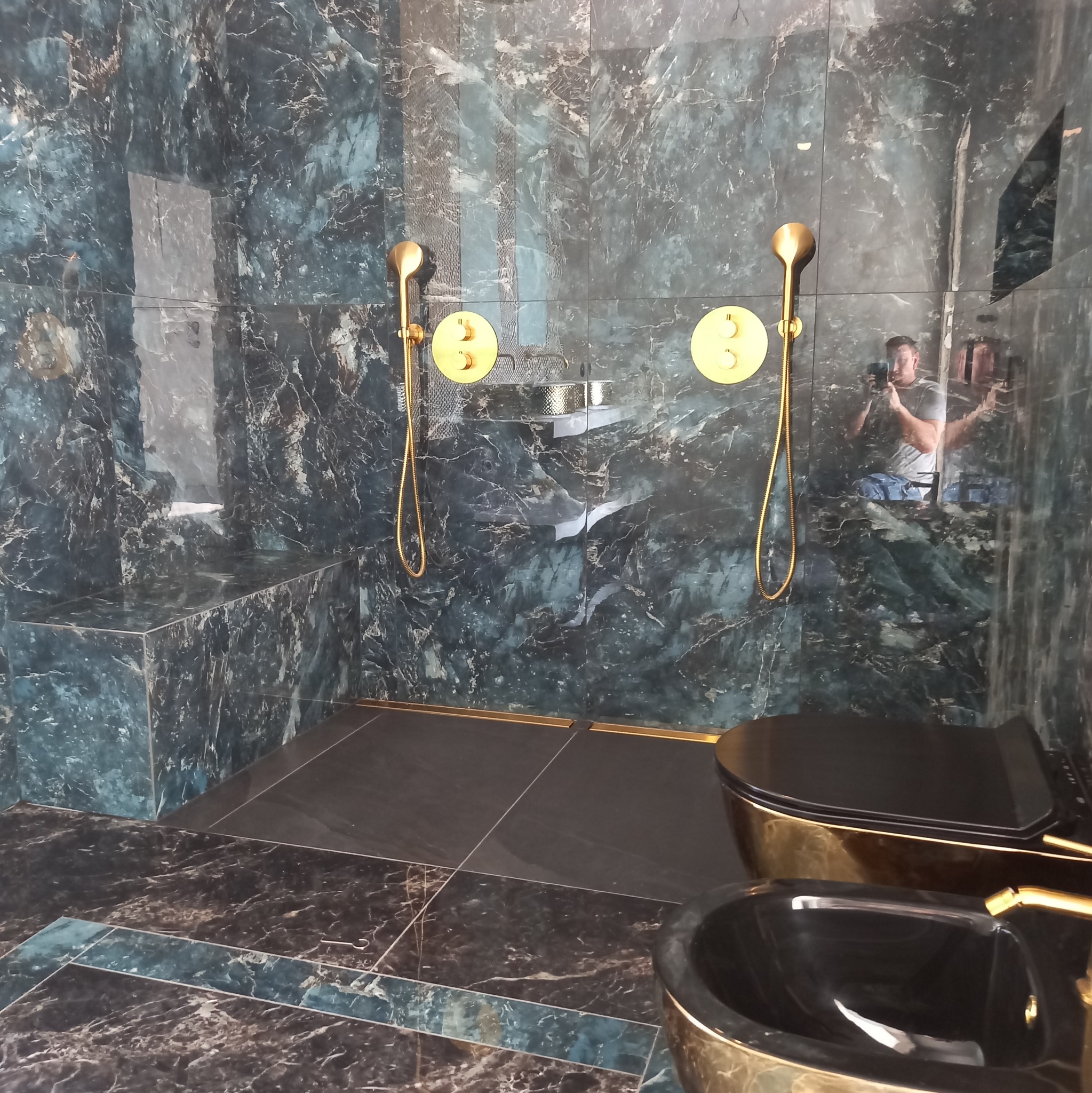 Luksusowa łazienka z ciemnymi marmurowymi płytkami, złotą armaturą i kabiną prysznicową typu walk-in. W lustrzanej powierzchni odbija się osoba robiąca zdjęcie.