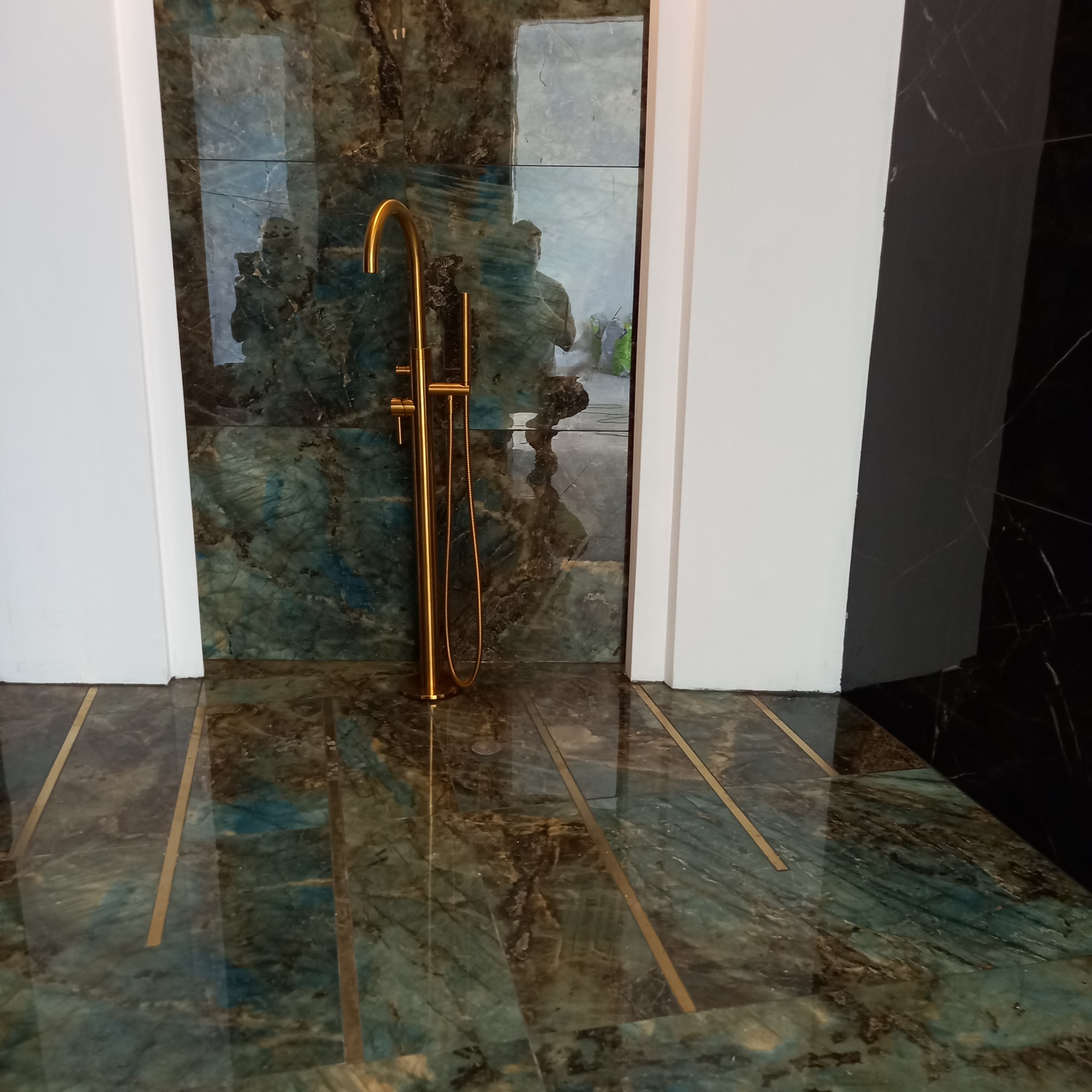 Błyszcząca, zielono-brązowa marmurowa ściana i podłoga z złotymi wstawkami, uzupełnione złotą armaturą prysznicową w nowoczesnej łazience.