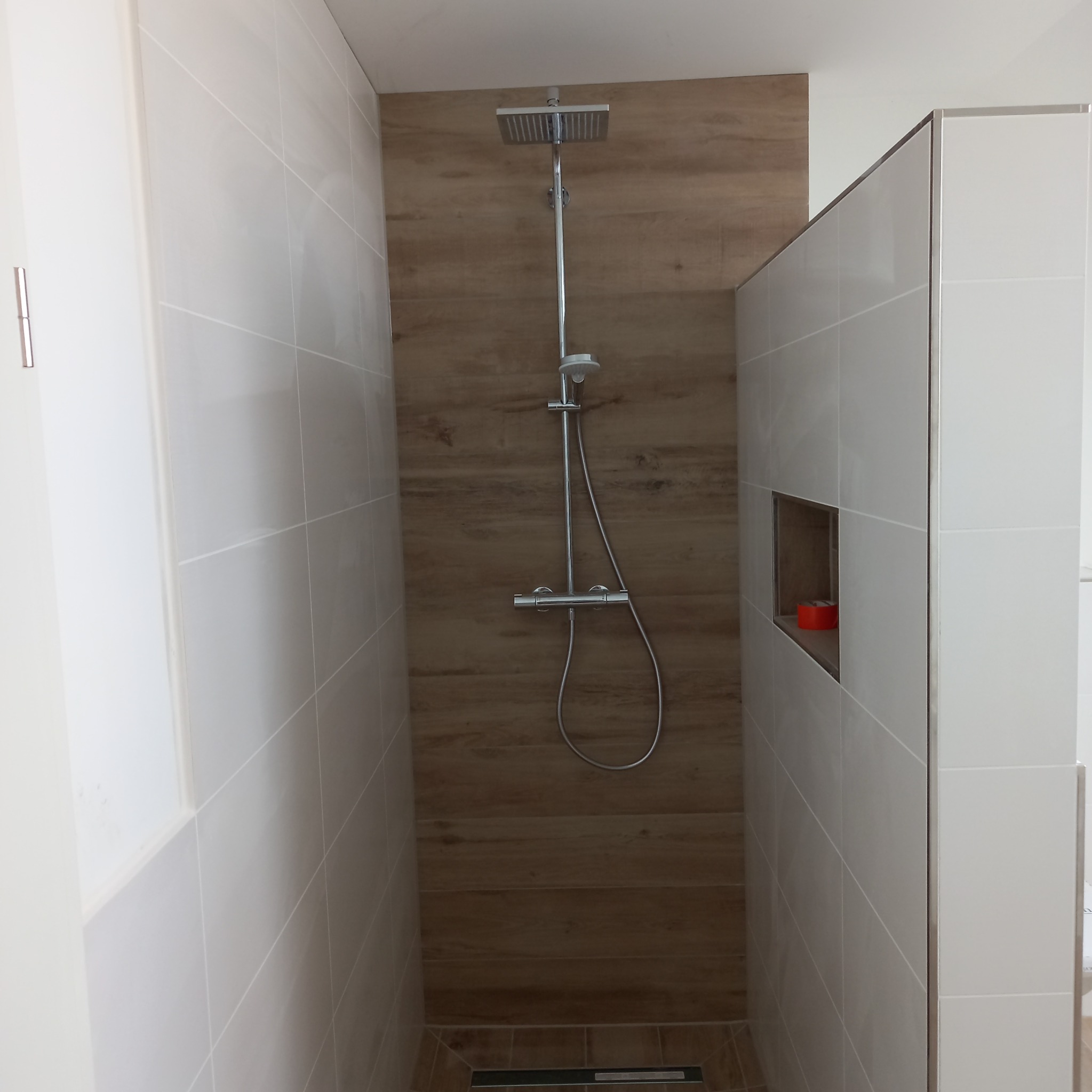 Nowoczesny prysznic typu walk-in z drewnopodobną ścianą i białymi płytkami, widoczny odpływ liniowy w podłodze.