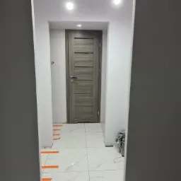 Korytarz po remoncie z białymi ścianami, białymi płytkami podłogowymi z szarymi żyłami, szarą stolarką drzwiową i pomarańczową taśmą malarską zabezpieczającą łączenia płytek.