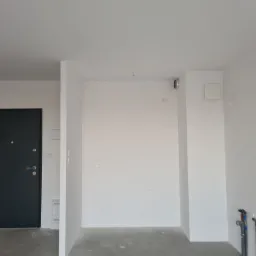 Wnętrze w stanie deweloperskim z gładkimi, białymi ścianami, czarnymi drzwiami wejściowymi z wizjerem i wystającymi rurami instalacji wodnej przy podłodze.