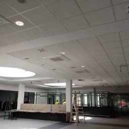 Multifinish - Sufit kasetonowy w jasnym, przestronnym pomieszczeniu biurowym z widocznymi kratkami wentylacyjnymi, oświetleniem punktowym i rozkładanymi schodami w tle.