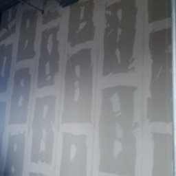 Multifinish - Ściana z płyt kartonowo-gipsowych w trakcie szpachlowania, widoczne łączenia płyt wypełnione masą szpachlową, metalowe profile konstrukcyjne przy ścianie, niebieska przesłona w tle.