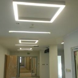 Multifinish - Korytarz biurowy z nowoczesnym oświetleniem LED w kształcie kwadratów i linii, białe ściany, szklane drzwi, znak ewakuacyjny.